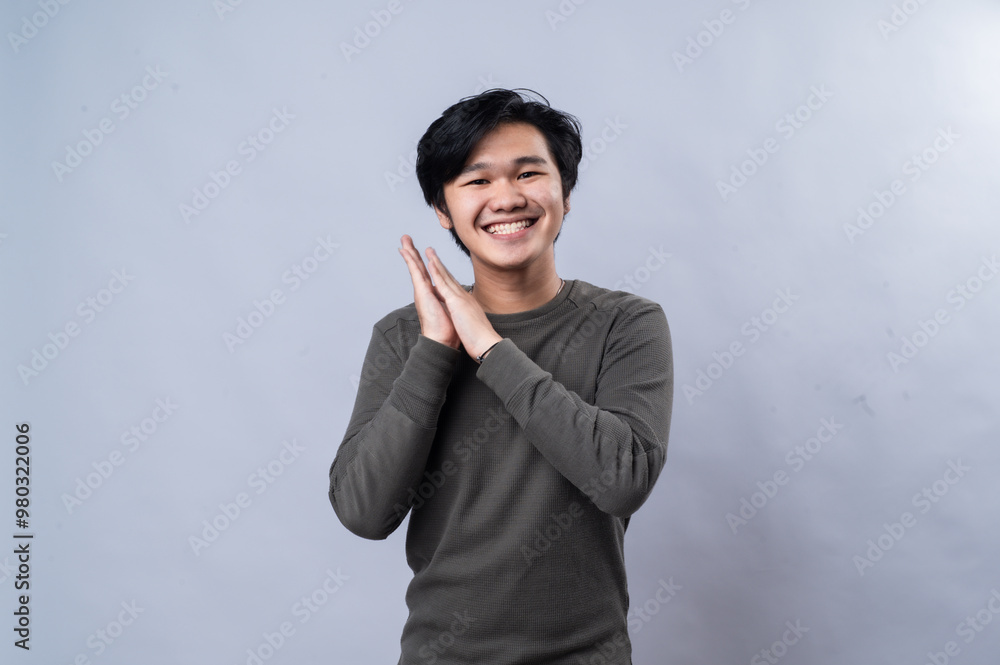 An Asian man wearing a long-sleeve gray shirt, looking excited and smiling while clasping his hands together. against a plain white background An Asian man wearing a long-sleeve gray shirt, looking excited and smiling while clasping his hands together. against a plain white background