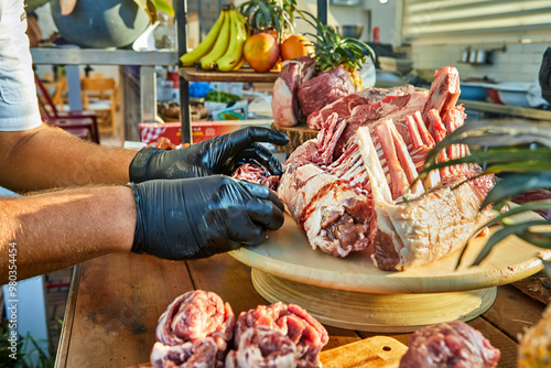 A chef wearing black gloves arranges a rack of raw lamb on a wooden board surrounded by various cuts of meat, preparing for cooking photo