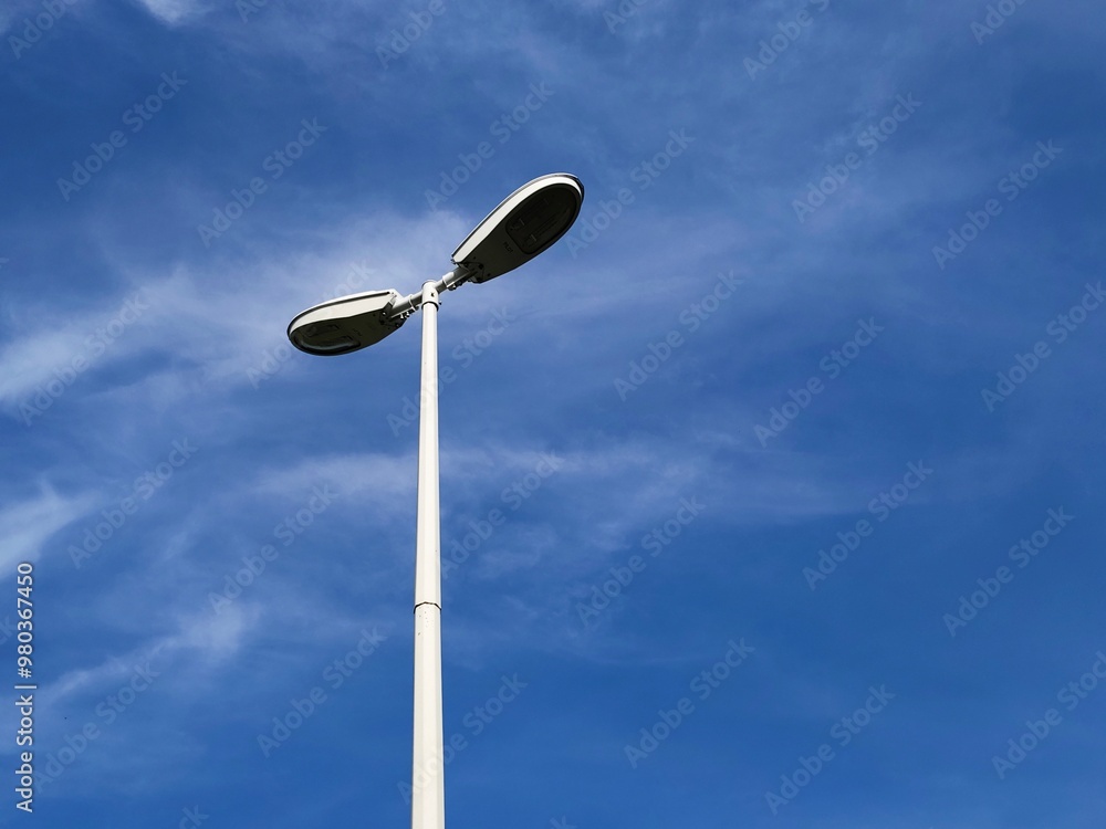 A modern street lamp with two lights, captured from a low angle against a bright blue sky with thin clouds. The lamp post stands in stark contrast to the clear sky A modern street lamp with two lights, captured from a low angle against a bright blue sky with thin clouds. The lamp post stands in stark contrast to the clear sky