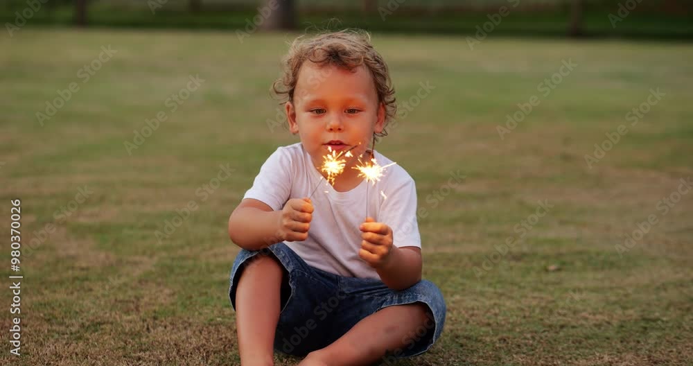 Cheerful young boy holds two glowing sparklers, fascinated by flickering lights. He watches them intently while talking, sparks flying in air. Slow motion shot with soft flickering glow Cheerful young boy holds two glowing sparklers, fascinated by flickering lights. He watches them intently while talking, sparks flying in air. Slow motion shot with soft flickering glow