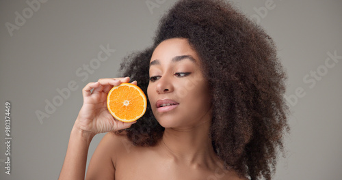 Beauty, vitamins and healthcare concept - beautiful African American woman with curly afro hairstyle and clean, healthy skin holding orange halves in her hands posing and looking at the camera