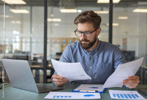 Stunned young office worker seated at a table, looking seriously surprised at the documents and bills he's holding.