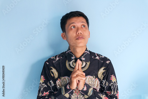 Asian young man wearing a batik shirt showing thinking something gesture with his eyes looking up photo