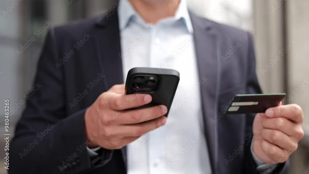 Close-up shot of a young businessman in a suit using his smartphone for online shopping. He is holding a credit card and entering the details on his phone. Close-up shot of a young businessman in a suit using his smartphone for online shopping. He is holding a credit card and entering the details on his phone.