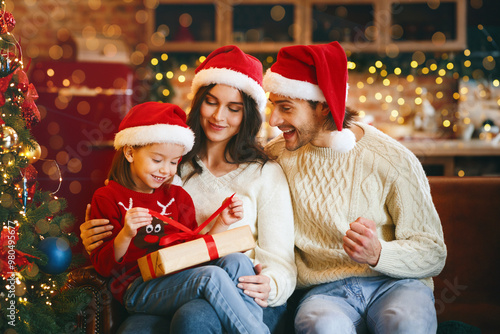 Cute little festive girl openng xmas gifts on Christmas eve with mom and dad, decorated kitchen interior, copy space