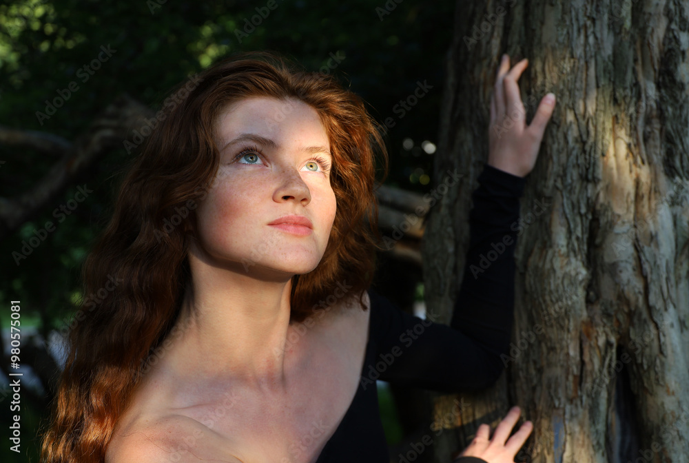 Naklejka premium Portrait of ginger girl with long red hair and freckles standing near the tree. Young sensual model, natural female beauty
