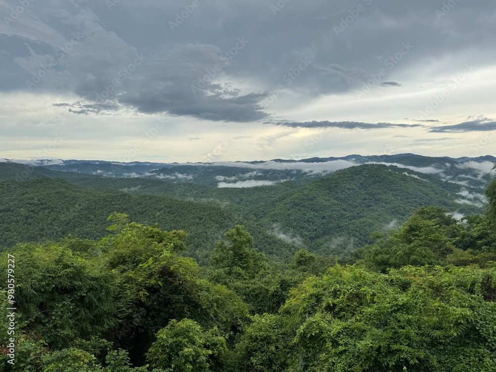 Obraz premium Expansive view of green mountains covered in mist, with dark clouds overhead creating a dramatic, atmospheric landscape. A perfect scene for themes of nature, wilderness, and adventure