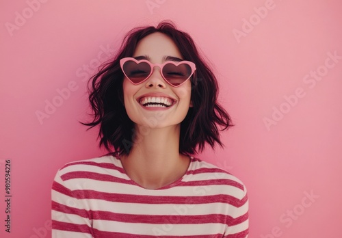 Portrait of a beautiful woman with a bob hairstyle, laughing and wearing heart-shaped sunglasses and a striped shirt on a pink background. Concept with copy space.