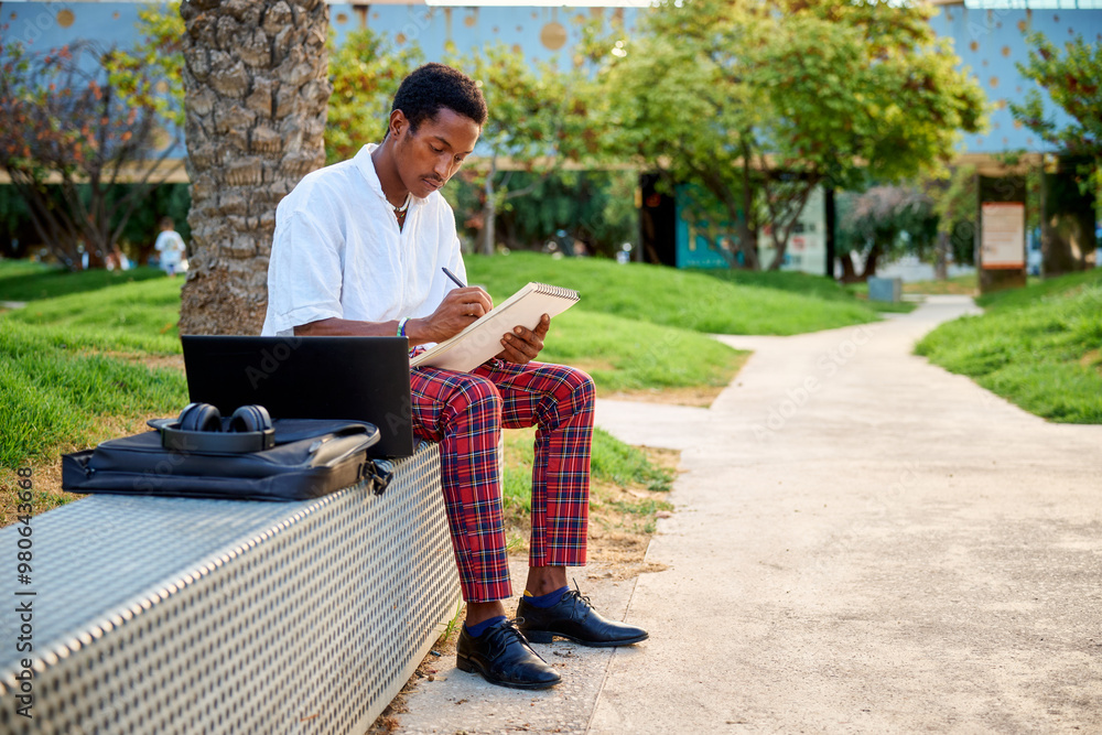 Fototapeta premium A young Black man sits on a bench in an urban area, sketching ideas in a notebook while his laptop and bag sit nearby