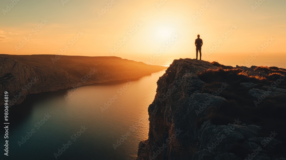 A silhouette of a person stands on a cliff at sunset, overlooking a serene body of water. A silhouette of a person stands on a cliff at sunset, overlooking a serene body of water.