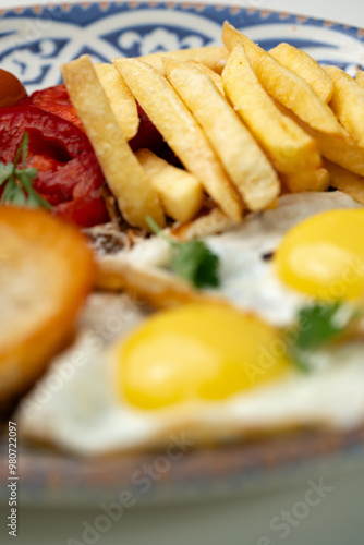 Breakfast plate with fried eggs, sausage, tomatoes, and fries served on a decorative blue plate