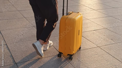Travel concept. Woman walks down street pushing her yellow suitcase on wheels in black trousers white sneakers . Outdoors in daylight, against smooth tiled surface. Suitcase bright yellow with rotate