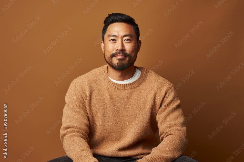 Obraz premium Portrait of a content asian man in his 30s wearing a cozy sweater while standing against soft brown background