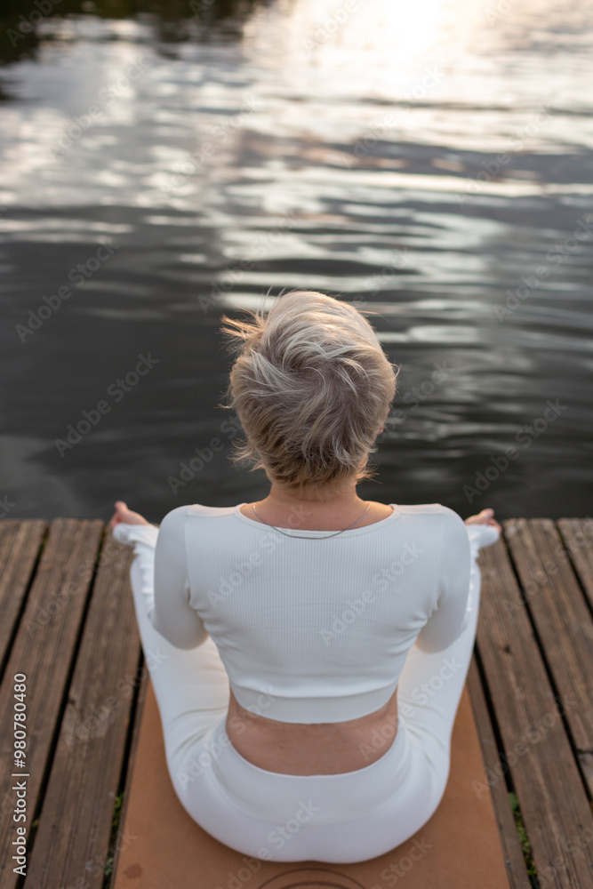Fototapeta premium Mature woman in a white suit in a lotus position on the river bank