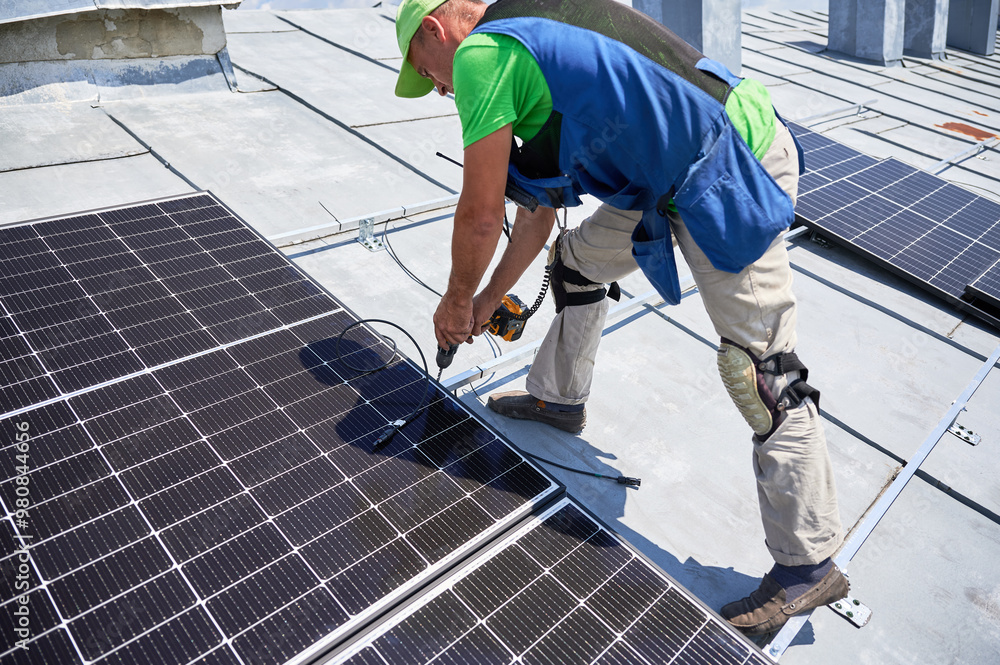 Worker building photovoltaic solar panel system on metal rooftop of house. Man technician installing solar module with help of electric drill outdoors. Alternative energy generation concept. Worker building photovoltaic solar panel system on metal rooftop of house. Man technician installing solar module with help of electric drill outdoors. Alternative energy generation concept.