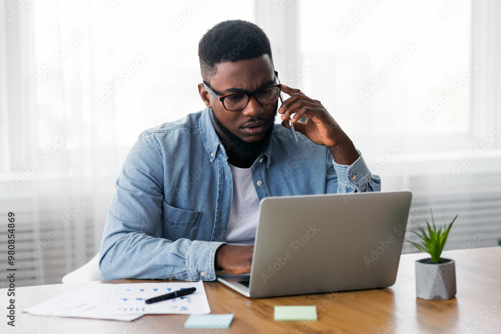 Concerned african american employee talking on mobile phone and looking at laptop screen, having issues with project, copy space Concerned african american employee talking on mobile phone and looking at laptop screen, having issues with project, copy space