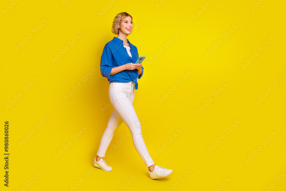 Full size portrait of pretty young lady hold phone walk empty space wear shirt isolated on yellow color background Full size portrait of pretty young lady hold phone walk empty space wear shirt isolated on yellow color background
