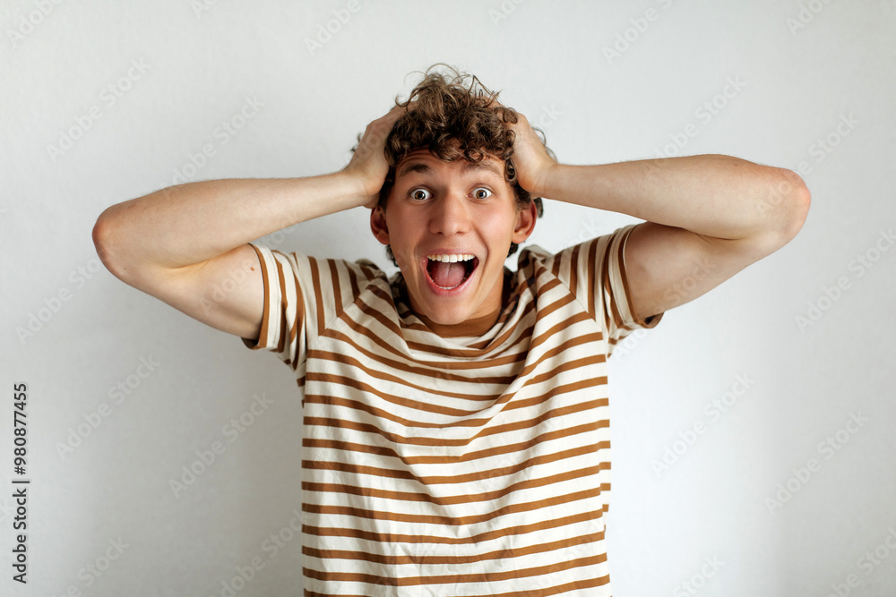 Young man expressing excitement with hands on his head in a simple indoor setting during daytime Young man expressing excitement with hands on his head in a simple indoor setting during daytime