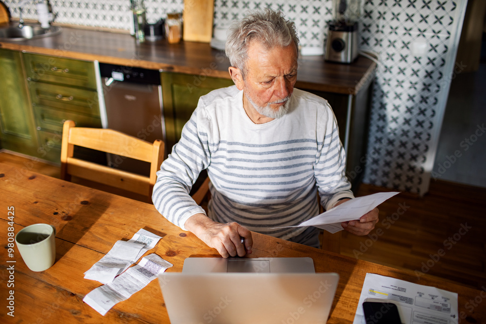 Elderly man managing bills and finances on laptop in kitchen Elderly man managing bills and finances on laptop in kitchen