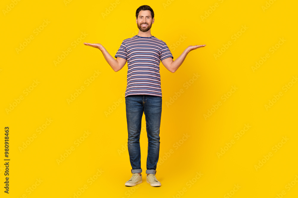 Full length photo of satisfied guy wear striped t-shirt jeans arms comparing products empty space isolated on yellow color background Full length photo of satisfied guy wear striped t-shirt jeans arms comparing products empty space isolated on yellow color background