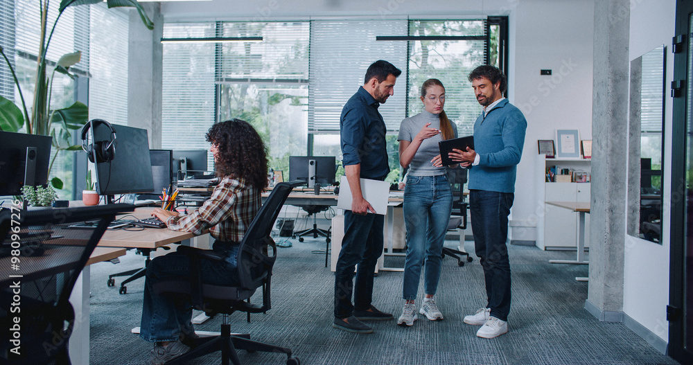 © ihorvsn - Casual day in office. People talking in middle of isle while looking at tablet device. Thinking about possible improvements. Woman on side putting on headset with microphone. Call center.