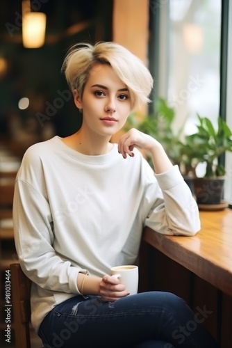 Blonde Woman in Cafe: A stylish young woman with a chic blonde pixie cut enjoys a moment of quiet contemplation in a cozy cafe, her relaxed pose and serene expression conveying a sense of peace and t