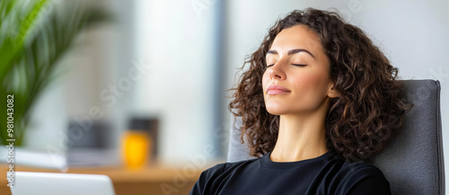 Profile shot of a person sitting in a chair, eyes closed and meditating, with an open laptop and office items softly blurred in the background