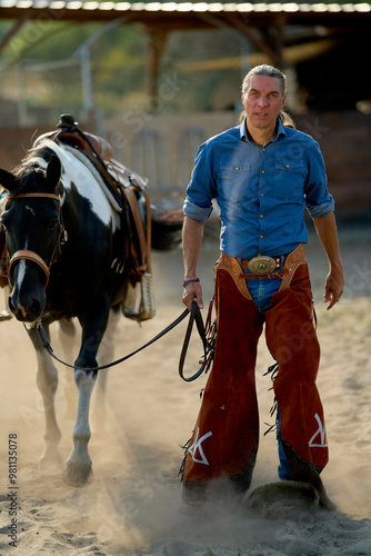 A confident man in cowboy attire walks beside a saddled horse in a dusty arena.