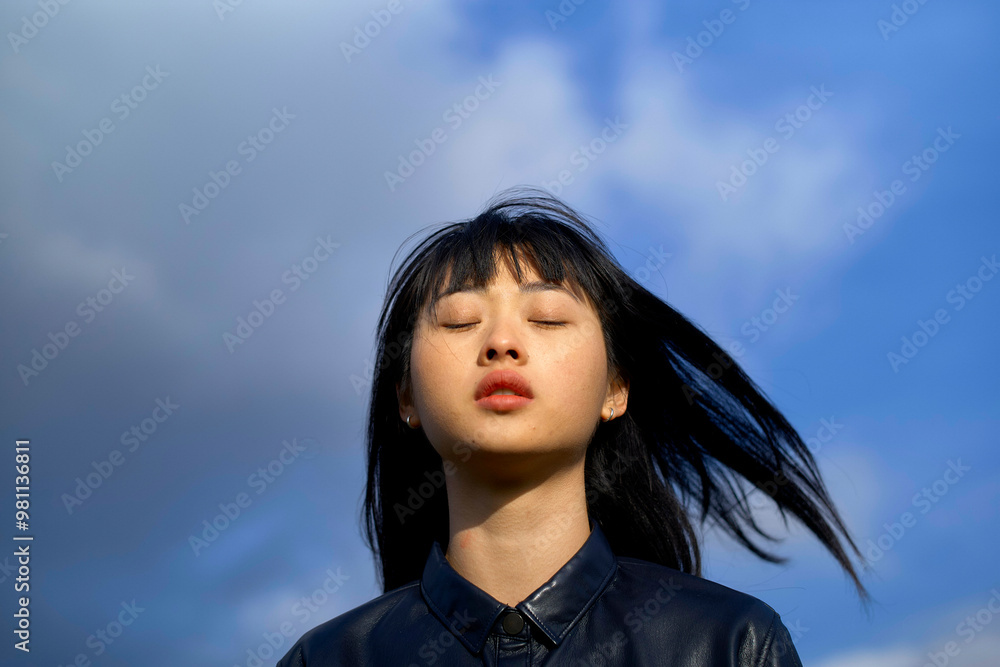 Close-up of a young woman with black hair and eyes closed against a blue sky with clouds. Close-up of a young woman with black hair and eyes closed against a blue sky with clouds.