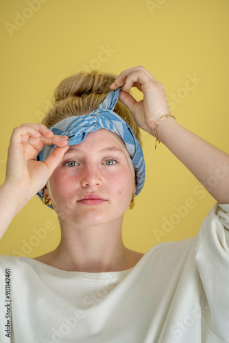 Young woman with blonde hair tying a blue headscarf against a yellow background, looking into the camera. photo