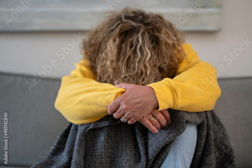 An adult with curly hair wearing a yellow sweatshirt and jeans sits hunched over on a grey couch, with their face obscured by their arms. photo