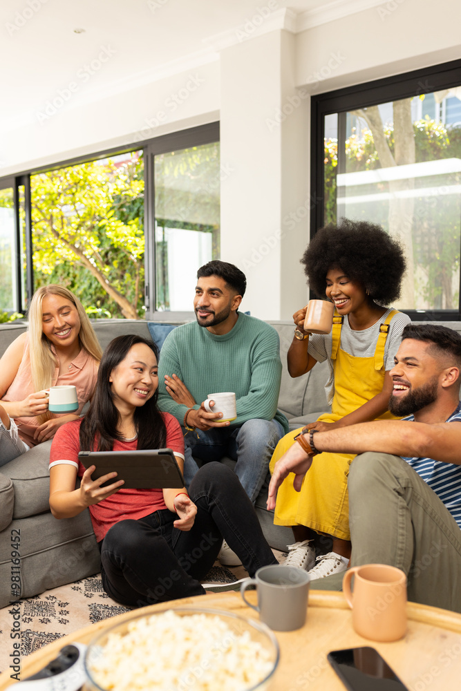 Diverse friends sitting on couch, drinking coffee, and watching tablet together, smiling Diverse friends sitting on couch, drinking coffee, and watching tablet together, smiling