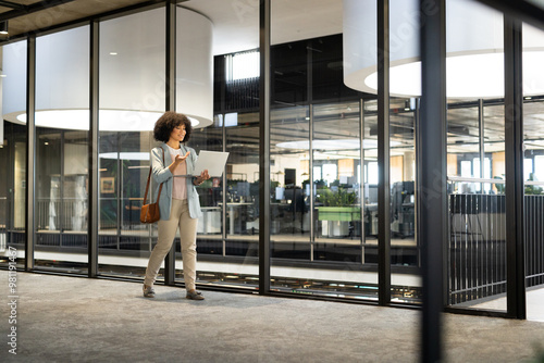 Walking in modern office, woman holding laptop and wearing business attire, copy space