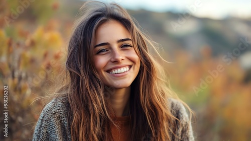 A joyful young woman with long hair smiles brightly in a natural outdoor setting, surrounded by autumn foliage.