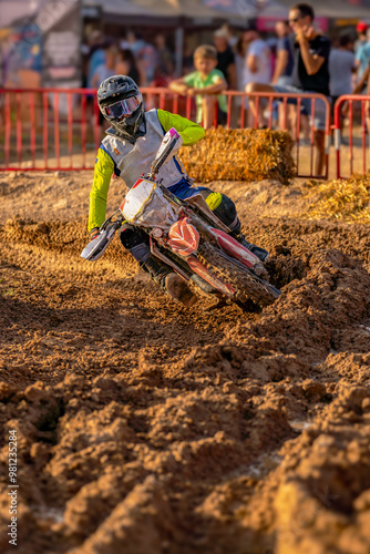 Motocross rider in action turning and folding his motorcycle while driving almost sinking on a muddy track, maintaining balance in a sharp curve while people watch the event behind protective fences. photo