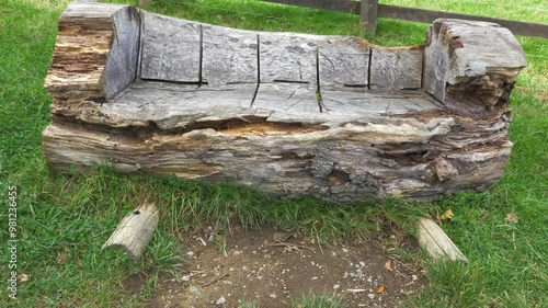 Closeup of an old weathered bench seat, which was roughly carved from a large tree trunk, positioned on grass next to a fence outside.