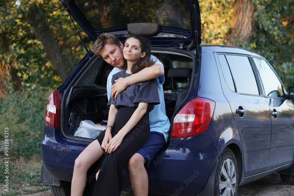 young couple sits together in the open trunk of a car parked in the forest. They are surrounded by trees and green vegetation young couple sits together in the open trunk of a car parked in the forest. They are surrounded by trees and green vegetation