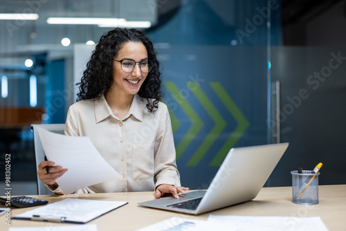 Smiling businesswoman working on laptop, reviewing papers in office. Professional setting reflects modern work environment. Ideal for concepts of productivity, technology, and corporate life.