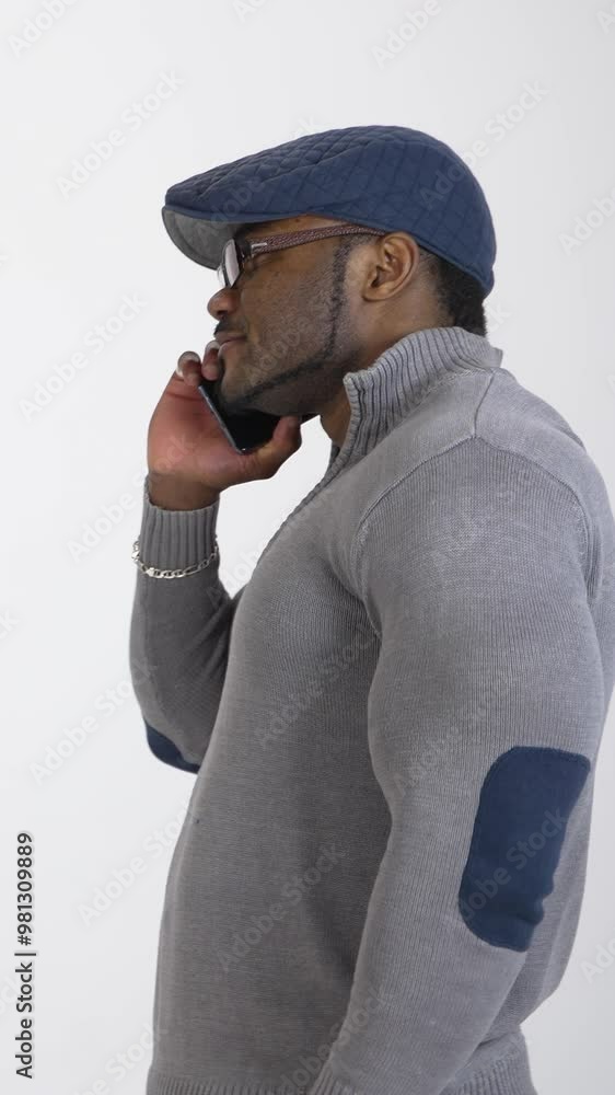 Serious black man with a phone in studio. Afro american guy in gray sweater and blue cap talking the mobile phone on white background. Side view. Vertical video Serious black man with a phone in studio. Afro american guy in gray sweater and blue cap talking the mobile phone on white background. Side view. Vertical video