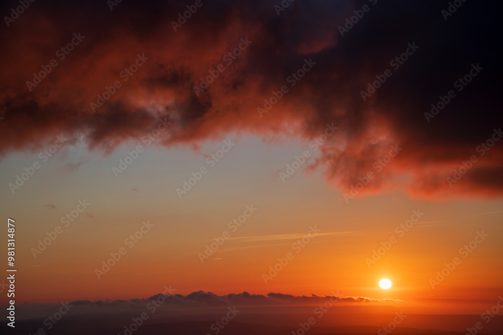 Fiery red and black clouds dominate a dramatic sunset sky, with the sun glowing on the right and a small patch of clear sky on the left, setting a striking backdrop Fiery red and black clouds dominate a dramatic sunset sky, with the sun glowing on the right and a small patch of clear sky on the left, setting a striking backdrop