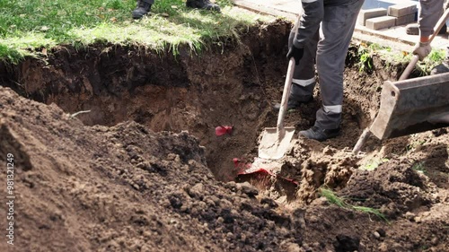 Teams are digging deep trench in residential yard, preparing for utility installation as they use shovels and machinery. Bright afternoon sun casts shadows on freshly turned earth, showing effort invo photo