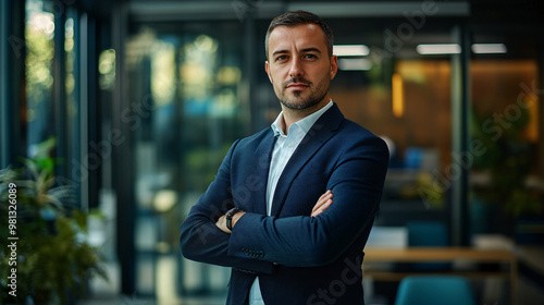 Businessman is standing in a modern office building with his arms crossed, looking serious and confident