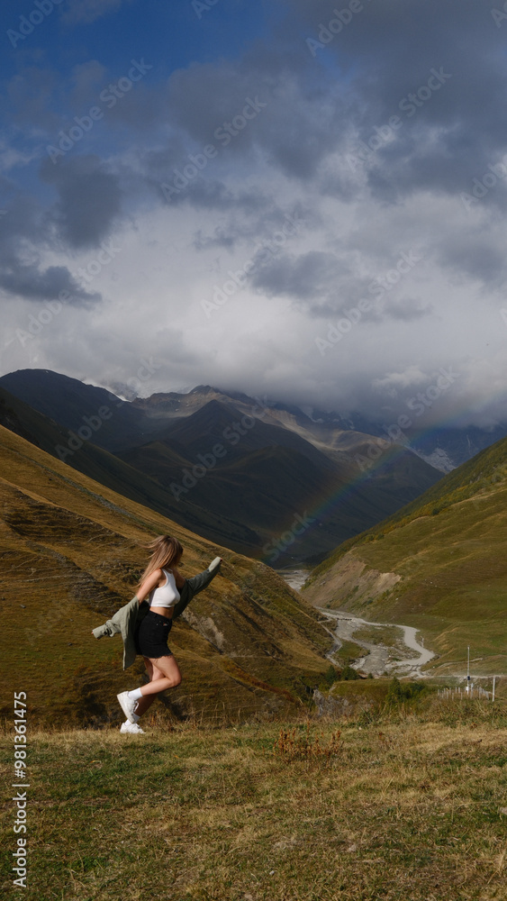 Naklejka premium A young woman leaps joyfully on a hillside, surrounded by vast mountains under a cloudy sky. A soft rainbow arcs in the background, adding a touch of magic to the dramatic scenery.