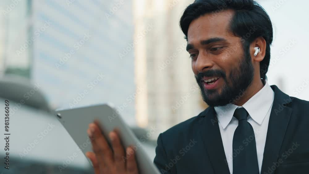 Portrait of handsome indian man in business suit waving greeting gesture while using digital tablet for video call. Friendly brunette using wireless earphones for better sound quality outdoors. Portrait of handsome indian man in business suit waving greeting gesture while using digital tablet for video call. Friendly brunette using wireless earphones for better sound quality outdoors.