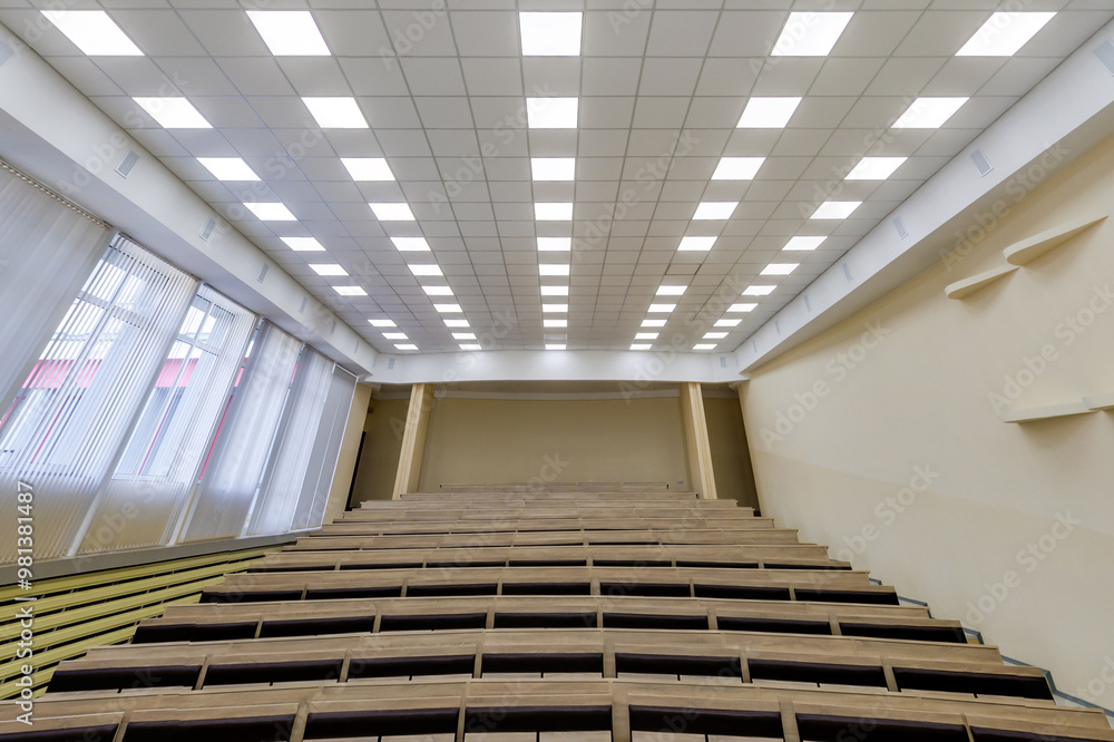 Obraz premium empty auditorium with tiered wooden seating and a bright ceiling featuring multiple square lights. Large windows with vertical blinds are on the left side, giving the room a clean, modern atmosphere