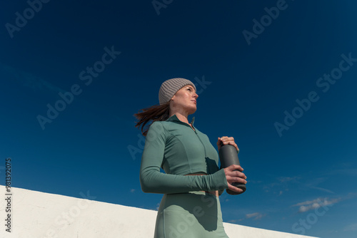 Canvas Print Sporty woman runner holding a aluminium water bottle, outdoors with blue sky bac