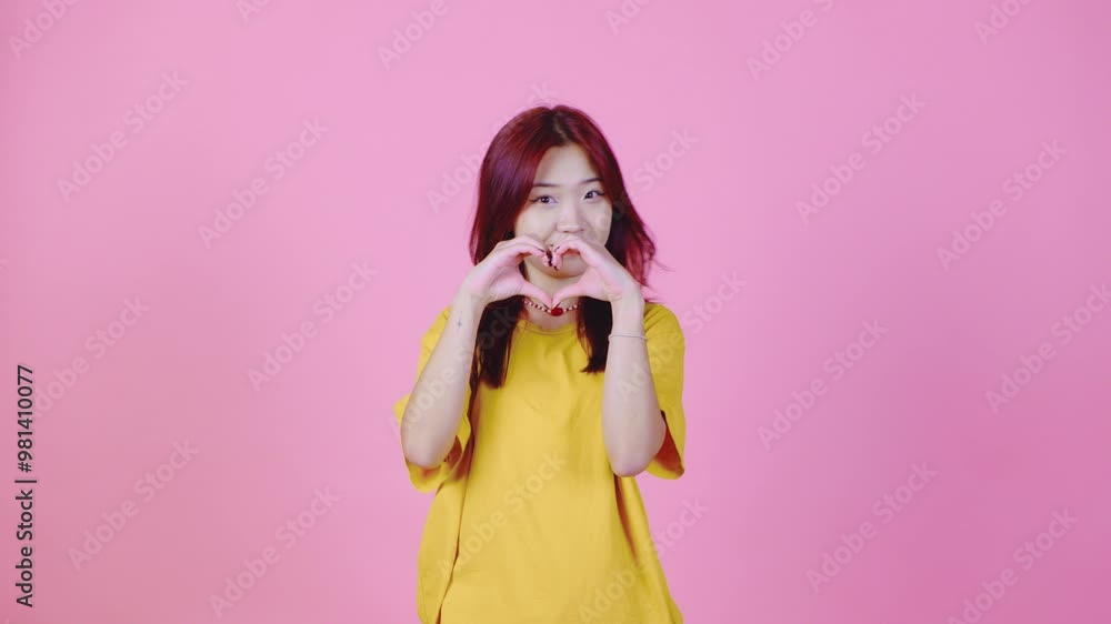 A young Asian woman with vibrant red hair stands against a pink background, forming a heart shape with her hands. Her sweet and loving expression adds warmth to the scene. A young Asian woman with vibrant red hair stands against a pink background, forming a heart shape with her hands. Her sweet and loving expression adds warmth to the scene.