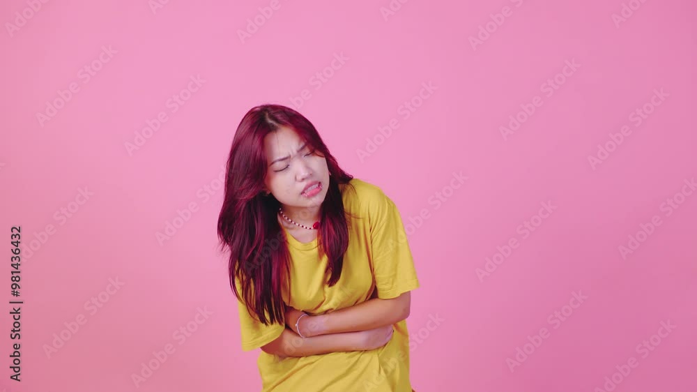 A young Asian woman with vibrant red hair, wearing a yellow t-shirt, stands against a pink background, holding her stomach with a pained expression. She appears unwell, struggling with discomfort A young Asian woman with vibrant red hair, wearing a yellow t-shirt, stands against a pink background, holding her stomach with a pained expression. She appears unwell, struggling with discomfort
