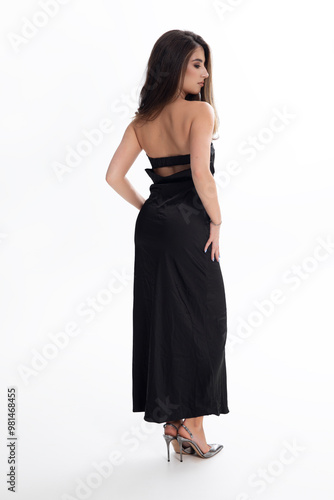 Back view of a young woman in elegant long dress and high heels, with makeup and hairstyle, over white background.