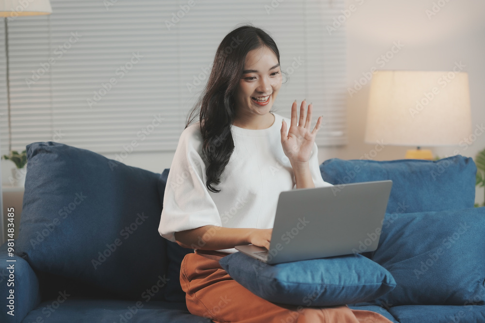 Fototapeta premium Young woman happily waves and smiles during a video call on her laptop in a cozy living room, embodying the concept of remote work and online communication